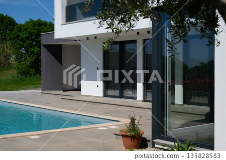 Modern luxury villa with swimming pool and clean minimalist architecture under a blue sky. Contemporary residential exterior featuring large windows, terrace, and elegant outdoor living space 135828583