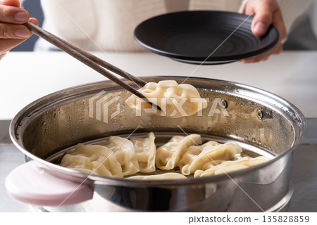 Hot steamed dumpling being picked up by chopsticks from a steamer pot to a black plate 135828859