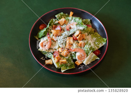 top view fresh Caesar salad with shrimp, croutons, tomatoes, and green vegetables in a ceramic black and red plate on green leather floor background top view fresh Caesar salad with shrimp, croutons, tomatoes, and green vegetables in a ceramic black and red plate on green leather floor background 135828920