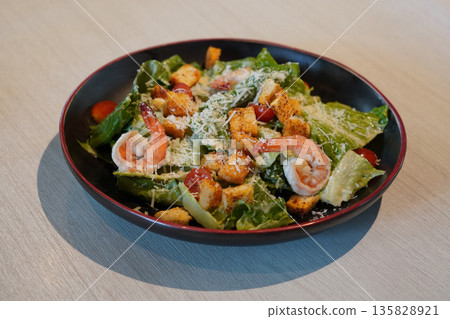 top view fresh Caesar salad with shrimp, croutons, tomatoes, and green vegetables in a ceramic black and red plate on wooden floor background top view fresh Caesar salad with shrimp, croutons, tomatoes, and green vegetables in a ceramic black and red plate on wooden floor background 135828921