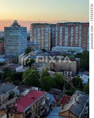 Cityscape. Old buildings with roofs, green trees, road and moving cars on sunny summer evening. Windows and walls of old houses, multi-story buildings in city. City scenery. Urban landscape. Cityscape. Old buildings with roofs, green trees, road and moving cars on sunny summer evening. Windows and walls of old houses, multi-story buildings in city. City scenery. Urban landscape. 135829316