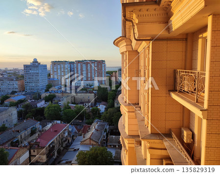 Cityscape with sea and blue sky on summer evening and sunlight illuminated white wall with windows of multi-story residential building. Old buildings, roofs, green trees. Front facade. Architecture 135829319