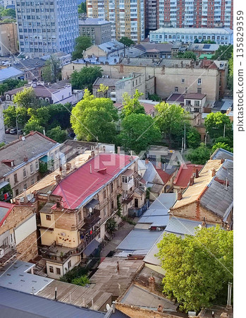 Cityscape. Old buildings with roofs, green trees, road and moving cars on sunny summer evening. Windows and walls of old houses, multi-story buildings in city. City scenery. Urban landscape. 135829359