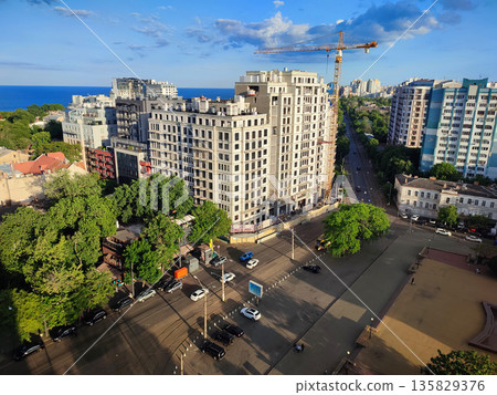 City street view from above. Large multi-story buildings, a square, green trees, an asphalt road for cars, moving cars and walking people on a sunny summer evening City street view from above. Large multi-story buildings, a square, green trees, an asphalt road for cars, moving cars and walking people on a sunny summer evening 135829376
