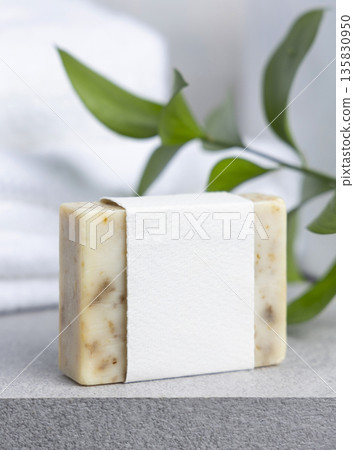 Soap bar with blank label against green leaves and white bath towels on grey stone, mockup 135830950