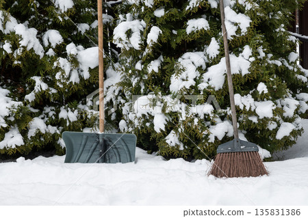 Snow shovel and broom standing in fresh snow near evergreen bushes. Winter household tools and seasonal outdoor maintenance concept. 135831386