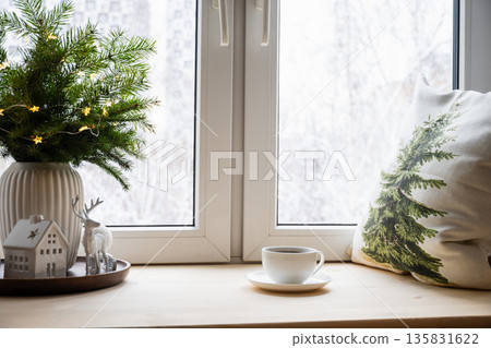 Cup of coffee on windowsill next to Christmas decorations fir branches fairy lights ceramic house and deer. Cozy winter interior. Festive holiday mood 135831622