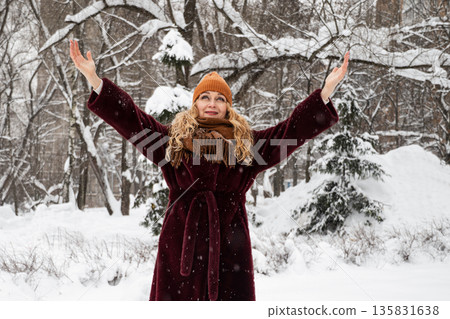 Smiling mature adult woman raising her arms and enjoying falling snow during winter walk in snowy city park. Female wearing warm coat, scarf and hat outdoors. Winter lifestyle, freedom, happiness and Smiling mature adult woman raising her arms and enjoying falling snow during winter walk in snowy city park. Female wearing warm coat, scarf and hat outdoors. Winter lifestyle, freedom, happiness and 135831638