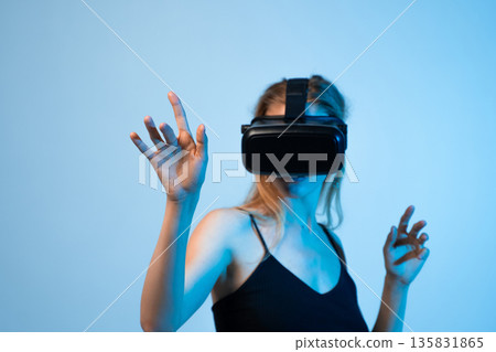 Young woman wearing virtual reality headset and interacting with digital interface using hand gestures in blue studio light 135831865