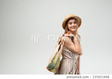 Smiling young woman in straw hat and summer dress carrying reusable mesh shopping bag with vegetables on light background Smiling young woman in straw hat and summer dress carrying reusable mesh shopping bag with vegetables on light background 135831888