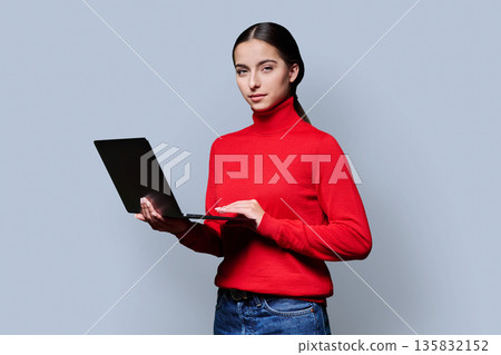 Portrait of serious young woman holding laptop on gray studio background Portrait of serious young woman holding laptop on gray studio background 135832152