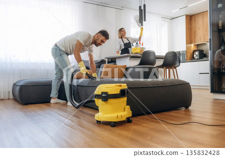 Modern yellow vacuum cleaner, busy. Two cleaning service workers are in domestic room 135832428