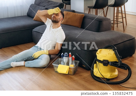 Tired, sitting on the floor and taking a break. Male professional cleaning service worker is domestic room Tired, sitting on the floor and taking a break. Male professional cleaning service worker is domestic room 135832475