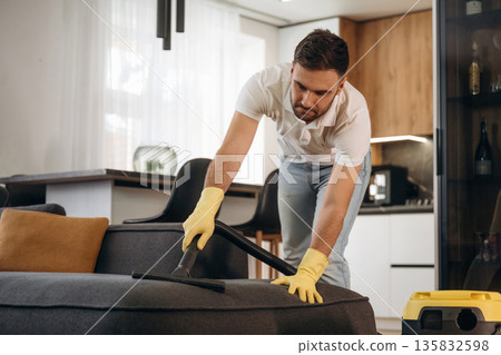 Care of sofa, using vacuum cleaner. Male professional cleaning service worker is domestic room Care of sofa, using vacuum cleaner. Male professional cleaning service worker is domestic room 135832598