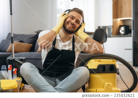 Enjoying music. Sitting. Male professional cleaning service worker is domestic room Enjoying music. Sitting. Male professional cleaning service worker is domestic room 135832621