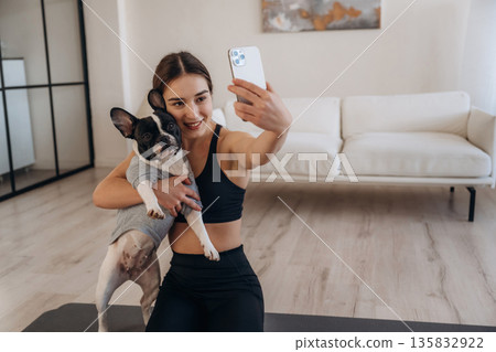 Doing selfie. Sportive woman in yoga clothes is with pug dog at home 135832922