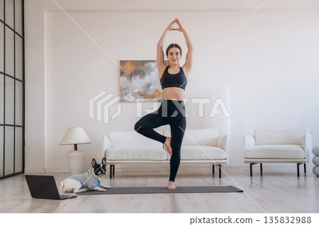 Tree exercise, standing on one leg. Woman with pug dog is doing yoga at home Tree exercise, standing on one leg. Woman with pug dog is doing yoga at home 135832988