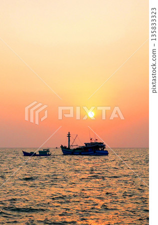 Sunset over ocean, casting orange pink colors in sky. Small fishing boats are visible in sea water Sunset over ocean, casting orange pink colors in sky. Small fishing boats are visible in sea water 135833323