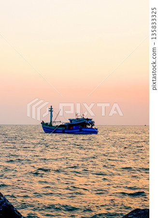 Sunset over ocean, casting orange pink colors in sky. Small fishing boats are visible in sea water 135833325