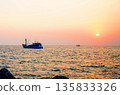 Sunset over ocean, casting orange pink colors in sky. Small fishing boats are visible in sea water 135833326