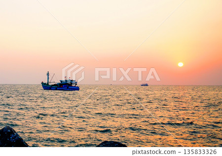 Sunset over ocean, casting orange pink colors in sky. Small fishing boats are visible in sea water 135833326