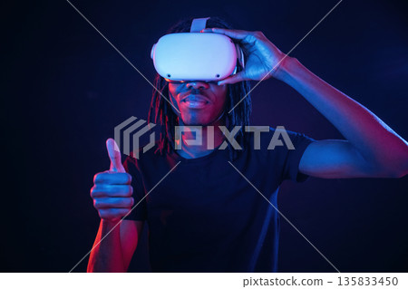 Thumb up gesture. African American guy is with virtual reality glasses in neon room 135833450