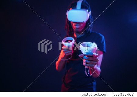 Controllers in hands. African American guy is with virtual reality glasses in neon room Controllers in hands. African American guy is with virtual reality glasses in neon room 135833468