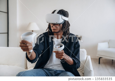 Relaxing by playing VR game, sitting. Black man in virtual reality glasses at home 135833535