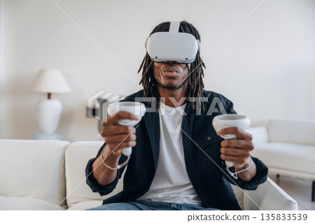 Sofa in domestic room, sitting with controllers. Black man in virtual reality glasses at home Sofa in domestic room, sitting with controllers. Black man in virtual reality glasses at home 135833539