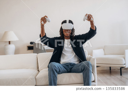 Celebrating victory in game. Black man in virtual reality glasses at home Celebrating victory in game. Black man in virtual reality glasses at home 135833586