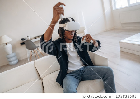 Hands up, celebrating victory. Black man in virtual reality glasses at home Hands up, celebrating victory. Black man in virtual reality glasses at home 135833595