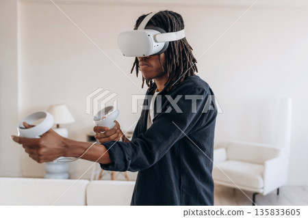 Shooting game, holding controllers. Black man in virtual reality glasses at home Shooting game, holding controllers. Black man in virtual reality glasses at home 135833605