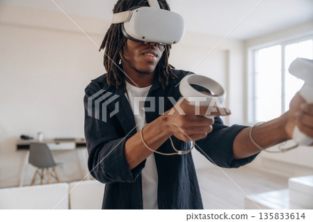 In casual clothes, headset and controllers. Black man in virtual reality glasses at home 135833614