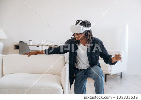 Hide and seek, near the sofa. Playing game. Black man in virtual reality glasses at home 135833639