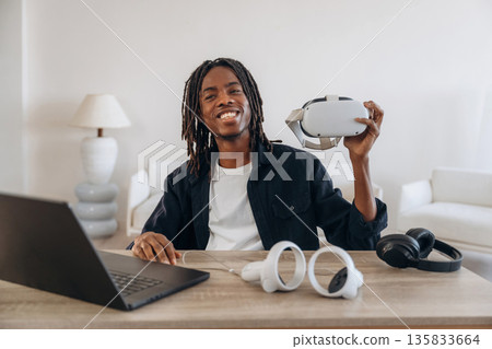 User is with headset. Young man is sitting by table with laptop and virtual reality glasses User is with headset. Young man is sitting by table with laptop and virtual reality glasses 135833664