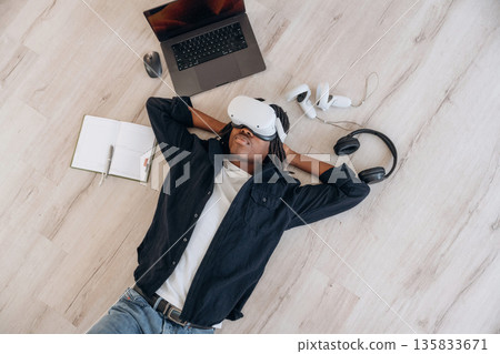 Laptop is nearby. Top view of man that is lying down on floor in virtual reality headset 135833671