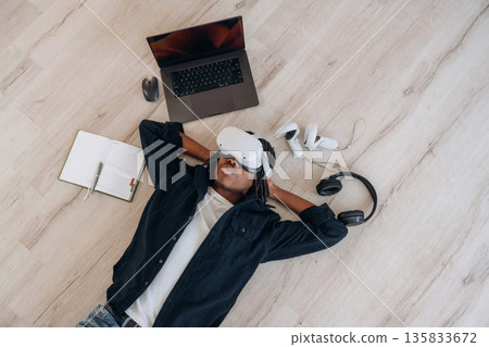 Laptop is nearby. Top view of man that is lying down on floor in virtual reality headset 135833672