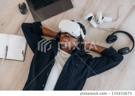 Laptop is nearby. Top view of man that is lying down on floor in virtual reality headset Laptop is nearby. Top view of man that is lying down on floor in virtual reality headset 135833673