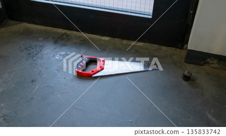 Hand saw lying on concrete floor in apartment building entrance. Tool for construction work or repair. Concept of home improvement and DIY. Hand saw lying on concrete floor in apartment building entrance. Tool for construction work or repair. Concept of home improvement and DIY. 135833742