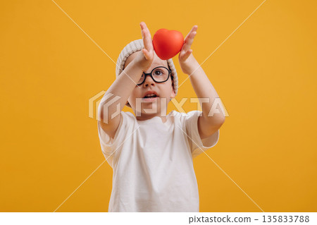 Red small heart toy in hands. Cute little boy is against yellow background 135833788