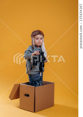 Black binoculars in hands, in aviator hat. Cute little boy is against yellow background Black binoculars in hands, in aviator hat. Cute little boy is against yellow background 135834167