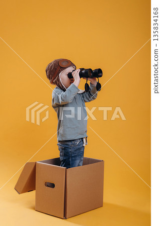 Black binoculars in hands, in aviator hat. Cute little boy is against yellow background Black binoculars in hands, in aviator hat. Cute little boy is against yellow background 135834168