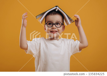 Book is on the head. Cute little boy is against yellow background Book is on the head. Cute little boy is against yellow background 135834225