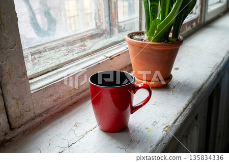Red mug on an old windowsill next to a houseplant, bringing comfort and warmth to an interior background. Old housing stock Red mug on an old windowsill next to a houseplant, bringing comfort and warmth to an interior background. Old housing stock 135834336
