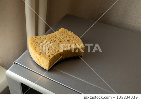 New sponge with black abrasive side on a grey microwave oven. Everyday kitchen cleaning and dishwashing tool for home hygiene. 135834339