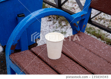 Paper cup on wet wooden bench during light hail or rain. Concept of cold weather, discarded items, urban scene. 135834343