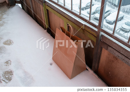 Brown paper delivery bag on snow covered balcony floor. food delivery service, eco friendly packaging, shopping Brown paper delivery bag on snow covered balcony floor. food delivery service, eco friendly packaging, shopping 135834355