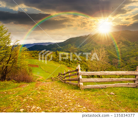 rolling hills of rural landscape in green grass at sunset. wooden fence in mountains in evening light. alpine countryside in spring. cloudy sky. hope, expectation, new beginning and bright future 135834377