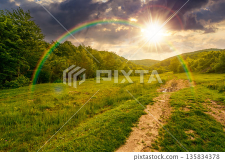 country dirt road hides in the woods at sunset. path through grassy alpine pasture in summer evening light. lovely countryside scenery in mountains. discover remote place for wanderlust under rainbow country dirt road hides in the woods at sunset. path through grassy alpine pasture in summer evening light. lovely countryside scenery in mountains. discover remote place for wanderlust under rainbow 135834378