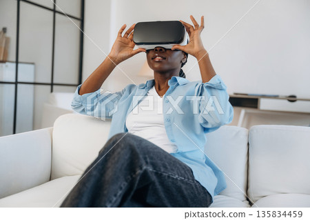 Augmented reality headset, sitting on sofa. Woman is at home in domestic room 135834459